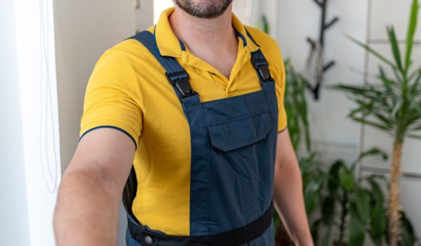 A cheerful handyman in a yellow shirt and blue overalls extends his hand for a handshake while holding a toolbox.